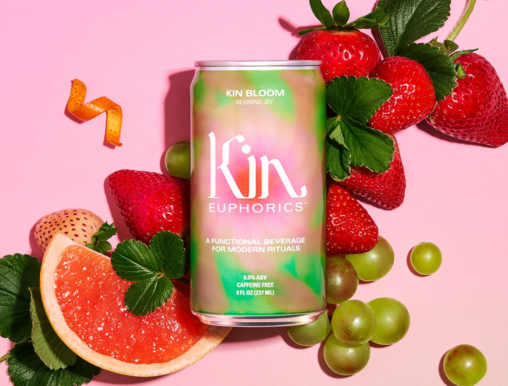 A can of Kin Euphorics Kin Bloom on a pink background with strawberries, grapefruit, white grape, orange peel and mint leaves