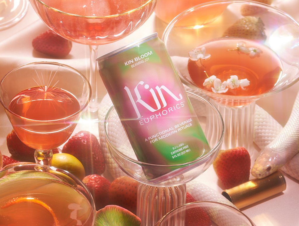A can of Kin Euphorics Kin Bloomin in champagne and wine glasses surrounded by a pink snake and red lipstick.