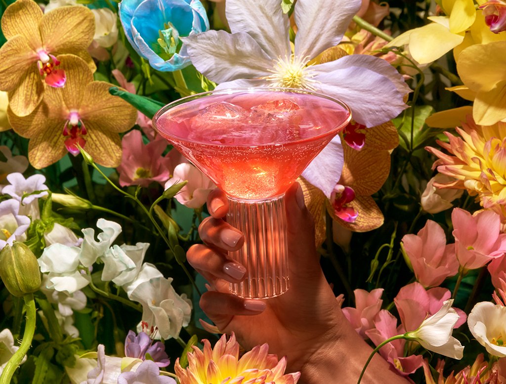 Hand holding a glass of Kin Euphorics Kin Bloom surrounded by flowers