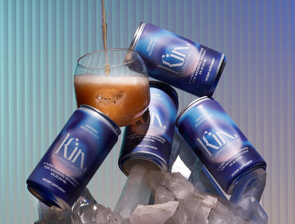 Multiple cans stacked of kin euphorics non-alcoholic calming beverage lightwave with one glass cup stacked on top of ice and crystals with liquid being poured in
